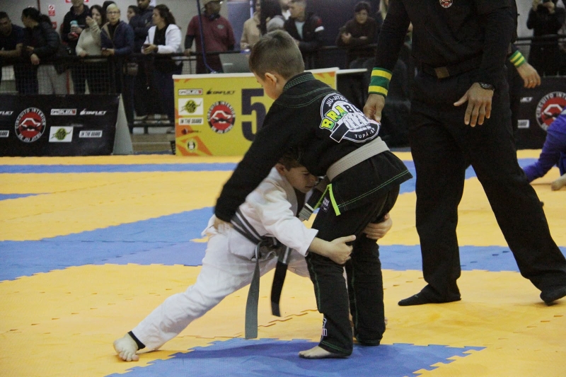 Atletas da categoria infantil disputam uma luta de jiu-jitsu em um tatame, acompanhados pelo árbitro.